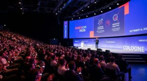 A keynote speaker presenting on stage at DMEXCO 2026 in Cologne, with a large screen displaying AI and digital marketing data behind them and a packed audience in the foreground
